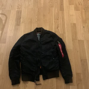 Svart bomberjacka från Alpha Industries - Säljer en klassisk svart bomberjacka från Alpha Industries i mycket bra skick. Jackan har en cool 'Remove Before Flight'-tag och är perfekt för höst och vår. Den är långärmad och har en dragkedja framtill. Insidan är i en kontrasterande orange färg som ger en snygg touch.