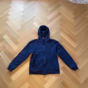 Mörkblå hoodie från Polo Ralph Lauren - Säljer en snygg mörkblå hoodie från Polo Ralph Lauren i storlek L/G dvs 14-16. Den har en dragkedja och en klassisk röd logga på bröstet. Perfekt klassisk tjocktröja för alla tillfällen. Säljer den nu endast för 399 då nypris ligger på runt 1200 kronor! Skriv gärna i DM om ni har några frågor! Mvh Sthlm garderoben.