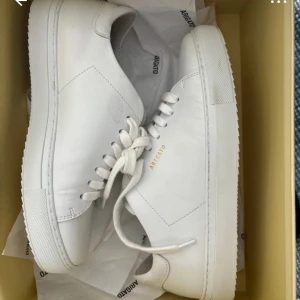 Vita sneakers från Axel Arigato - Säljer ett par stilrena vita sneakers från Axel Arigato i nytt skick. Skorna är i skinn med klassisk snörning och en diskret logga i guld på sidan. Perfekta för både vardag och fest!
