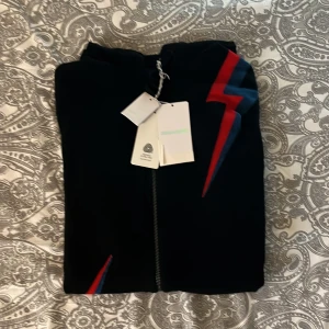 Svart hoodie med blixtdetaljer från Zadig & Voltaire - Säljer en svart hoodie från Zadig & Voltaire i nytt skick. Den har en cool blixtdetalj i rött och blått på framsidan. Perfekt för höst och vår, med dragkedja och huva för extra stil och komfort. Passar både till vardags och festliga tillfällen!