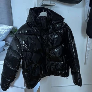 Svart glansig pufferjacka - Säljer en svart glansig pufferjacka i mycket bra skick. Jackan har en dragkedja framtill och är perfekt för kalla dagar. Den är EU-storlek 38 och har en mysig, vadderad design som håller dig varm och stilren.