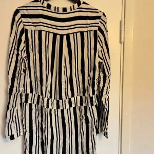 Randig klänning med knytband - Säljer en superfin svartvit randig klänning med långa ärmar. Den har ett knytband i midjan för en snygg passform. Perfekt för både vardag och festliga tillfällen. Klänningen är i bra skick och redo för en ny ägare!