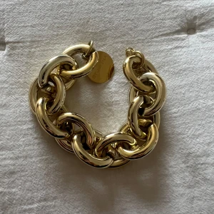 Guldarmband från By Jolima - Armband Bonnie från By Jolima, Snyggt och trendigt guldarmband med stora, glänsande länkar. Perfekt för att ge en lyxig touch till din outfit. Armbandet har en justerbar kedja för perfekt passform och en rund platta som detalj. Passar både till vardag och fest!