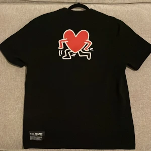 Svart t-shirt från Axel Arigato - Säljer en svart t-shirt från Axel Arigato med ett coolt rött hjärtmotiv på baksidan. Framsidan har texten 'ARIGATO CO2014' och 'Keith Haring' i rött. Perfekt för en casual look eller fest. T-shirten är i bra skick och gjord av mjuk bomull.