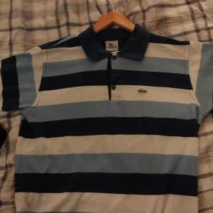 Randig pikétröja från Lacoste - Säljer en snygg randig pikétröja från Lacoste i olika blå nyanser. Tröjan har en klassisk krage och knappar framtill. Perfekt för en avslappnad men stilren look. Passar både till vardags och lite finare tillfällen.