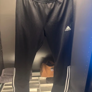 Svarta träningsbyxor från Adidas - Snygga svarta träningsbyxor från Adidas med klassiska vita ränder längs sidorna. Byxorna har en elastisk midja och är perfekta för sport och träning. Adidas-loggan är tryckt på framsidan.