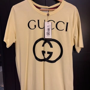 Gul t-shirt från Gucci - Säljer en gul t-shirt från Gucci med det ikoniska logotyptrycket i svart på framsidan. T-shirten har en rund halsringning och korta ärmar. Perfekt för en stilren och lyxig look, skick 10/10. Priset kan diskuteras