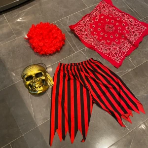 Randig piratkostym med dödskallemask - Cool piratkostym från Marks & Spencer för barn i åldern 7-8 år. Inkluderar randiga byxor i rött och svart, en guldig dödskallemask och en röd bandana. Perfekt för maskerad eller Halloween!