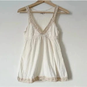 Vit ärmlös topp med spetsdetaljer - Säljer en söt vit ärmlös topp med v-ringning och spetsdetaljer vid hals och nederkant. Perfekt för varma dagar eller som ett lager under en kofta. Toppen har en luftig passform och är gjord i ett mjukt material. Storlek M (S)