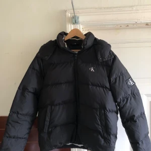 Svart pufferjacka från Calvin Klein Jeans - Säljer en svart pufferjacka från Calvin Klein Jeans. Jackan har en dragkedja framtill och en huva för extra värme. Den är dekorerad med CK-logotypen på bröstet och ärmen. Perfekt för kyliga dagar.