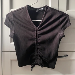 Svart ribbad topp - Snygg svart ribbad topp med korta ärmar och v-ringning. Toppen har en knytning framtill som ger en fin detalj. Perfekt för en stilren look.