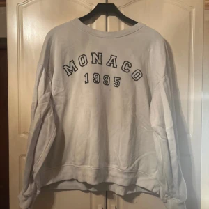 Vit sweatshirt från H&M med tryck - Säljer en vit sweatshirt från H&M med texten 'MONACO 1995' tryckt på framsidan. Tröjan har en avslappnad passform och långa ärmar, perfekt för en casual look. Den är gjord i ett mjukt material som känns skönt mot huden.