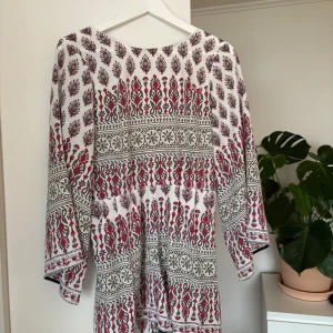 Vit och röd mönstrad byxdress - Säljer en vit byxdress med rött och grönt mönster. Byxdress har långa ärmar och ett unikt, detaljerat mönster som ger en bohemisk känsla. Perfekt för en avslappnad stil.