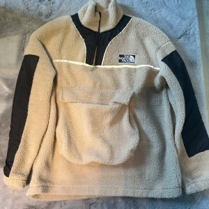 Beige fleecejacka från The North Face - Säljer en mysig beige fleecejacka från The North Face med svarta detaljer på ärmarna och en halv dragkedja framtill. Jackan har en stor ficka på magen och är perfekt för kyliga dagar. Den har en avslappnad passform och är långärmad.