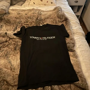 Svart t-shirt från Tommy Hilfiger - Snygg svart t-shirt från Tommy Hilfiger med klassisk logga på bröstet. Perfekt för en stilren look. T-shirten är kortärmad och gjord i mjukt material.