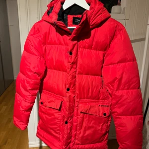 Röd dunjacka från Everest - Säljer en röd dunjacka från Everest med huva och knäppning framtill. Jackan har praktiska fickor med knappar och är perfekt för kalla dagar. Den är både stilren och funktionell, vilket gör den till ett bra val för vintern.