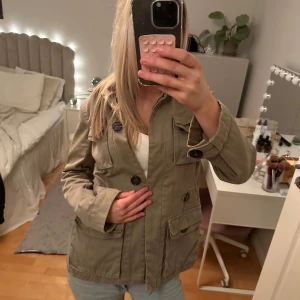 Militärgrön jacka med knappar - Säljer en snygg militärgrön jacka med stora knappar och flera fickor framtill. Jackan har en normal passform och långa ärmar, perfekt för vår och höst. En stilren och praktisk jacka för alla tillfällen.