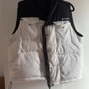 Vit och svart dunväst från FB Sister - Säljer en snygg vit och svart dunväst från FB Sister med dragkedja och knappar. Västen har två fickor framtill och märket 'Endlessly' broderat på bröstet. Perfekt för kyligare dagar!