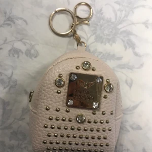 Beige miniryggsäck med nitar - Söt beige miniryggsäck med guldfärgade nitar och en metallplatta framtill. Den har en praktisk nyckelring och är perfekt som accessoar. Tillverkad i syntetmaterial med en texturerad yta. Helt ny 