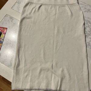 Vit kjol från Indiska - Snygg vit kjol från Indiska i storlek XL. Kjolen har en enkel design med dekorativa svarta stygn längs fållen. Perfekt för en stilren look.