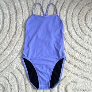 Lila baddräkt från Jolyn - Snygg lila baddräkt från Jolyn med korsade axelband och svart foder. Perfekt för simträning eller en dag på stranden. Tillverkad i 100% polyester med återvunnet foder för extra komfort och hållbarhet.