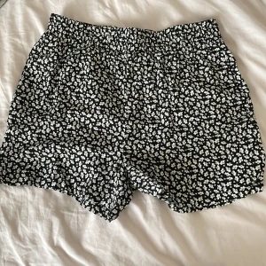 Svarta blommiga shorts från XOXO Friends - Snygga svarta shorts med ett vitt blommönster från XOXO Friends. Perfekta för en avslappnad stil med elastisk midja för extra komfort. Passar utmärkt till varma dagar och ger en trendig look.