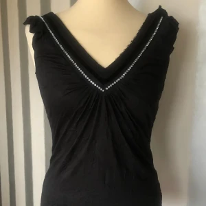 Svart ärmlös topp med pärldetaljer - Elegant svart ärmlös topp med v-ringning och pärldetaljer längs halsringningen. Toppen har en smickrande passform och är perfekt för en stilren look.