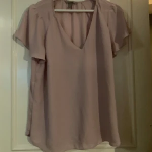Ljusrosa blus med v-ringning - Säljer en ljusrosa blus med v-ringning och korta ärmar. Blusen har en luftig passform och är perfekt för varmare dagar. Den är enkel och stilren, vilket gör den lätt att matcha med olika outfits.