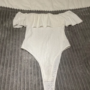 Vit offshoulder bodysuit - Säljer en stilren vit bodysuit med offshoulder-design och volangdetaljer. Perfekt för en somrig look. Materialet är lätt och luftigt med ett diskret mönster. 