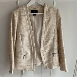 Beige/guld-kavaj från H&M - Beige kavaj med guldtrådar från H&M i storlek 40. Kavajen har en elegant struktur med dragkedjor vid fickorna och vid ärmslut. Figursydd. 