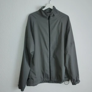 Grå vindjacka från Nike Golf - Säljer en grå vindjacka från Nike Golf med Storm-Fit teknologi. Jackan har en dragkedja framtill och elastiska ärmslut för extra komfort. Perfekt för blåsiga dagar på golfbanan eller andra utomhusaktiviteter.