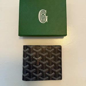 Plånbok Blå  - Snygg och stilren plånbok från G*ya**med deras ikoniska mönster. Plånboken har flera fack för kort och sedlar, perfekt för att hålla ordning på dina tillhörigheter. Kommer i en elegant grön förpackning.