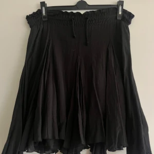 Svart blus med volangdetaljer - Snygg svart blus med volangdetaljer och elastisk axlar. Perfekt för en stilren look. Kjolen har en luftig passform och är lätt att matcha med olika toppar. Axellös