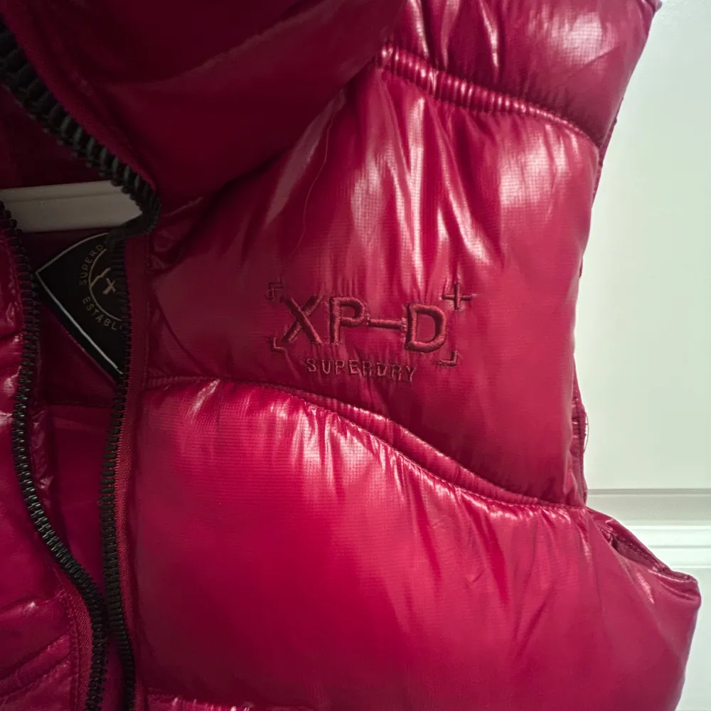 Säljer en snygg röd dunväst från Superdry med huva och dragkedja. Perfekt för kyliga dagar när du vill hålla värmen utan att kompromissa med stilen. Västen har en cool XP-D logga på bröstet.. Takit.