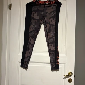 Camo mönstrade tights - Snygga camo mönstrade tights med svarta sidopaneler och orange detaljer i midjan. Perfekta för träning med en tight passform som ger bra stöd. Elastisk midja för extra komfort. Det är två stycken par i det här paketet men de ena paret är hel svarta istället för camo. Och de är samma storlek på båda par.