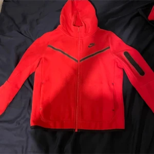 Röd Nike tech set. Kan säljas separat! - Säljer en snygg röd Nike tech set med dragkedja och huva. Den har svarta detaljer och en ficka på ärmen. Perfekt för en sportig look eller avslappnad stil. 