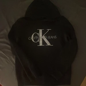 Svart hoodie från Calvin Klein Jeans - Säljer en stilren svart hoodie från Calvin Klein Jeans med det ikoniska CK-logotypen i vitt på ryggen. Perfekt för en avslappnad look. Hör av dig vid intresse!