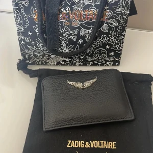 Svart plånbok från Zadig & Voltaire - Snygg svart plånbok i skinn från Zadig & Voltaire med ett coolt vingmotiv i silver på framsidan. Perfekt för att hålla dina kort och kontanter organiserade. Stilren och praktisk design som passar alla tillfällen.