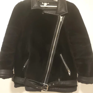 Svart pälsjacka med dragkedja - Säljer en svart pälsjacka med skinnkrage och sned dragkedja. Jackan har två fickor med dragkedjor och ett bälte i nederkant. Perfekt för kyliga dagar och ger en cool look.