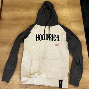 Grå och vit hoodie från Hoodrich - Säljer en snygg grå och vit hoodie från Hoodrich med svart text på bröstet. Hoodien har en stor ficka framtill och justerbar huva med dragsko. Perfekt för en avslappnad stil.