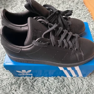 Svarta Adidas Stan Smith sneakers - Snygga och stilrena svarta Adidas Stan Smith sneakers i skinn. Skorna har klassisk snörning och en diskret design som passar till många olika stilar. Perfekta för dig som gillar en minimalistisk look.