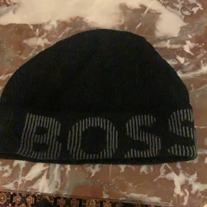 Svart mössa från Boss - Snygg svart mössa från Boss med stor logga framtill. Tillverkad i ett mjukt och bekvämt material som passar perfekt för kyligare dagar. Stilren design som funkar till det mesta.