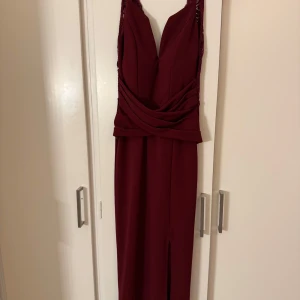 Vinröd balklänning med spetsdetaljer - Elegant vinröd långklänning med spetsdetaljer och djup v-ringning. Klänningen har en smickrande omlott-design i midjan och en slits framtill. Perfekt för festliga tillfällen.