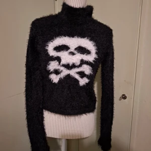 Svart fluffig polotröja med dödskallemotiv - Säljer en svart fluffig polotröja från Current Mood med ett stort vitt dödskallemotiv på framsidan. Tröjan har långa ärmar och en hög polokrage, perfekt för att hålla sig varm och stilren. Passar perfekt för en edgy look! Obs den är mindre i storlek passar från s till xl 
