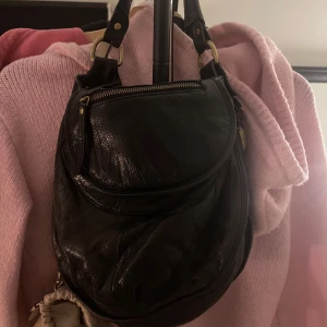 Svart axelväska i skinn - Snygg svart axelväska i skinn med dragkedja och praktiska fack. Perfekt för att bära med sig det viktigaste. Stilren design med justerbara remmar och metallspännen. Köpt på en secondhand i newyork, vet tyvärr inte märket!