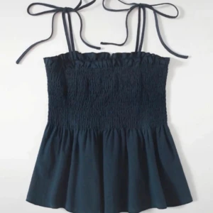 Svart axelbandslös topp med knytband - Snygg svart axelbandslös topp med knytband vid axlarna. Toppen har en smockad överdel och en luftig nederdel som ger en fin passform. Perfekt för varma dagar!