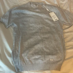 Grå kortärmad tröja från SAMS - Snygg grå tröja från SAMS med korta ärmar och ett diskret rutmönster. Tröjan har en rund halsringning och en liten SAMS-logga på framsidan. Perfekt för en stilren look.