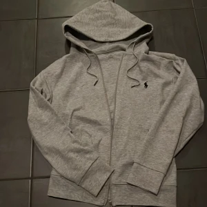 Grå hoodie från Ralph Lauren - Säljer en stilren grå hoodie från Ralph Lauren med dragkedja och justerbar huva. Perfekt för en avslappnad look. Den har en liten logotyp broderad på bröstet. Passar bra till både jeans och joggers.