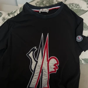 Svart t-shirt från Moncler - Snygg svart t-shirt från Moncler med ett stort, stilrent logotryck i vitt och rött på framsidan. T-shirten har korta ärmar och en rund halsringning. Perfekt för en casual look. Endast använd fåtal gånger 😊