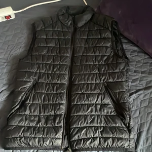 Svart quiltad dunväst - Säljer en stilren svart quiltad dunväst med dragkedja. Perfekt för kyligare dagar och enkel att matcha med olika outfits. Västen har en hög krage och två sidofickor med dragkedja.